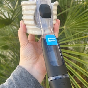 May include: A black and clear plastic dish soap dispenser with a white brush attached. The dispenser has a label that says "Soap on Demand".