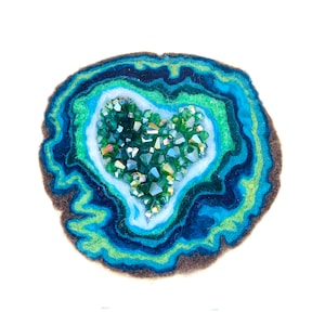 May include: A felt geode with a heart-shaped crystal cluster in the center. The geode is made of layers of blue, green, and brown felt, and the crystals are a sparkly green.
