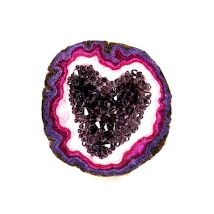 May include: A purple heart-shaped crystal made of small beads is set inside a round, layered felt geode. The geode has layers of pink, white, purple, and brown felt.