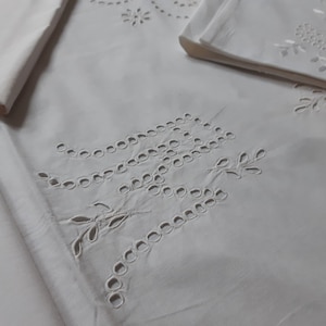 Op de afbeelding: Wit katoenen stof met delicate geborduurde bloemenmotieven. De stof heeft een subtiele glans en een zachte textuur.