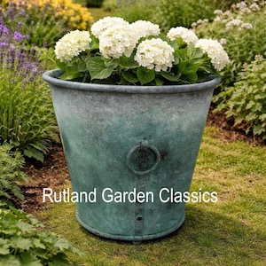 May include: A large, weathered, teal metal planter filled with white hydrangea flowers. The planter has a ring handle and is set in a garden with green grass and other plants. Text on the planter reads "Rutland Garden Classics."