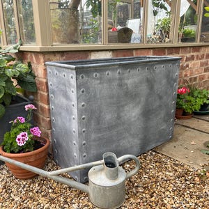 May include: A large, rectangular galvanised metal planter with riveted details, suitable for outdoor use. The planter is positioned near a brick wall and a greenhouse, with a watering can in the foreground. Various potted plants and flowers are visible.