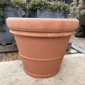 May include: A large terracotta planter with a wide, rounded base and a slightly flared top. The planter has a smooth, reddish-brown finish.