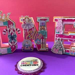 May include: A decorative "ONE" display with a Nutcracker theme. The letters are pink, gold, and white, featuring a crown, candy cane, ballerina, snow globe, Christmas tree, and drum. The name "Ellie" is also included.