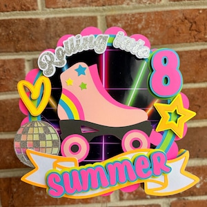 May include: A layered paper cake topper featuring a pink roller skate, a silver glitter disco ball, and a yellow heart and star. The text reads Rolling into 8 Summer in bright pink, blue, and silver glitter lettering against a black grid background.