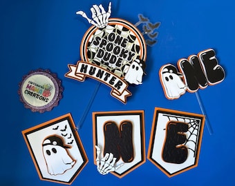 May include: Halloween-themed cake toppers and banner set. The set includes a round topper with the text "ONE SPOOKY DUDE HUNTER", a "ONE" topper, and a banner spelling "ONE". The toppers feature ghosts and bats.