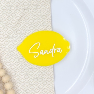 Puede incluir: Una tarjeta de lugar de forma de limón amarilla con el nombre "Sandra" escrito en letra cursiva blanca.