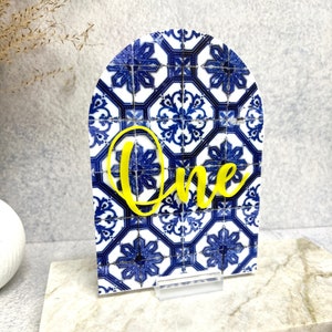 May include: A blue and white patterned arch-shaped table number with the word "One" in yellow lettering.