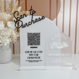 May include: White acrylic sign with a black cursive inscription "Scan to Purchase". A QR code is displayed with the text "venmo" below it. The sign also includes the text "SCAN THE QR CODE WITH YOUR CAMERA PHONE OR CONTACT HOST FOR OTHER PAYMENT OPTIONS". A second white acrylic sign with a semi-circular arch shape features the text "PLEASE VISIT WWW.THEHIDDENVUE.COM FOR DIRECT BOOKING AND MORE!"