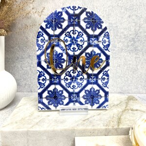 May include: Blue and white patterned arch-shaped table number with the word "One" in gold lettering.