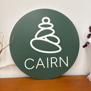 Peut inclure: Un panneau circulaire vert foncé présentant un graphique en ligne blanche représentant un cairn de pierres empilées, situé au-dessus du mot CAIRN en lettres capitales blanches. Le design est épuré avec des éléments blancs en relief.