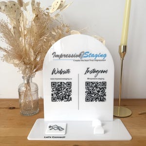 Può includere: Un cartello in acrilico bianco a forma di arco, con il testo "Impressive Staging" e lo slogan "Create the best first impression". Il cartello include codici QR per il sito web "www.impressivestaging.ca" e l'account Instagram "@impressive_staging". Il cartello ha anche un pulsante con il testo "Let's Connect!" e una grafica di un telefono con un simbolo di tocco.