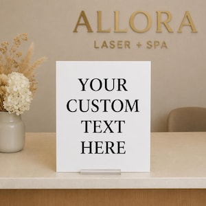 May include: A square white sign with a clear acrylic stand featuring the black printed text YOUR CUSTOM TEXT HERE. The sign is positioned on a light-coloured counter next to a ceramic vase filled with dried pampas grass and white hydrangea flowers.