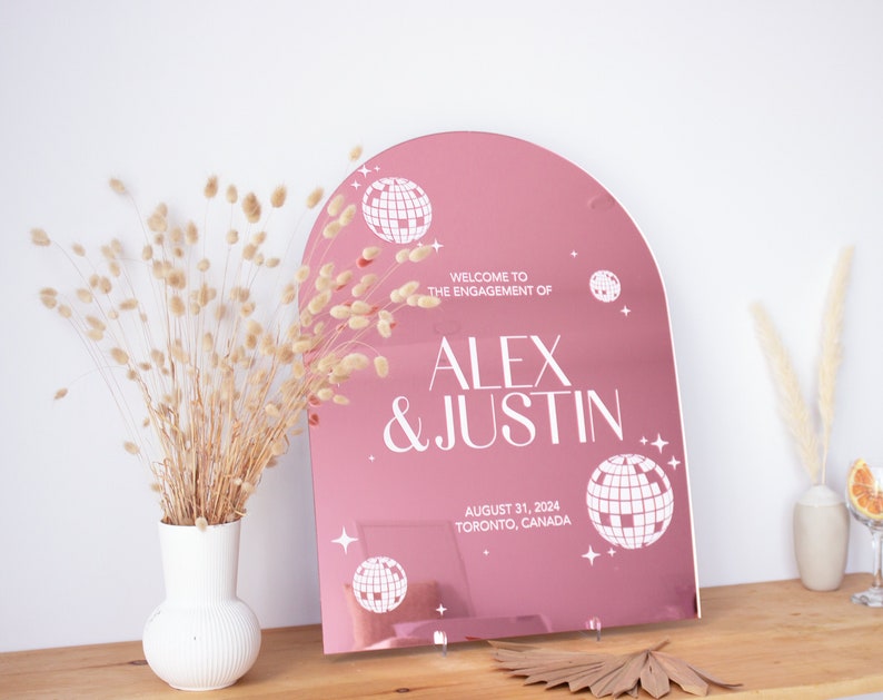 May include: Pink mirrored arch sign with white text and disco ball graphics. The text reads "Welcome to the engagement of Alex & Justin, August 31, 2024, Toronto, Canada."