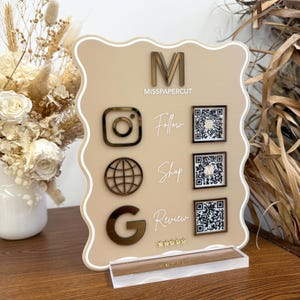 Op de afbeelding: Een beige acrylbord met een golvende rand, met de letter "M" en de tekst "MISSPAPERCUT". Het bevat pictogrammen voor Instagram, een wereldbol en Google, samen met QR-codes om te volgen, te winkelen en te beoordelen. Het bord is gemonteerd op een heldere acryl basis.