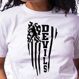 Op de afbeelding: Wit T-shirt met een zwarte afbeelding van een honkballer die een bal slaat voor een Amerikaanse vlag met het woord "DEVILS" er verticaal naast geschreven.