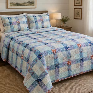 May include: A quilt and two matching pillow shams featuring a patchwork design of blue, light blue, white, and lavender squares. The fabric displays various floral and geometric patterns, creating a grid-like appearance across the bedding set.