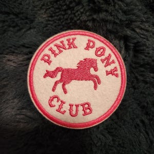 Peut inclure: Un écusson circulaire avec un fond blanc et une bordure rose. L'écusson présente les mots "PINK PONY CLUB" en lettres roses, entourant une silhouette rose d'un cheval.