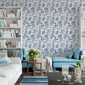 K&ouml;nnte beinhalten: Ein Wohnzimmer mit einem wei&szlig;en Sofa, blauen und wei&szlig;en Kissen mit Mustern, einem blauen Sofa und einem wei&szlig;en B&uuml;cherregal voller B&uuml;cher. Die Wand ist mit einer blauen und wei&szlig;en Blumentapete bedeckt.