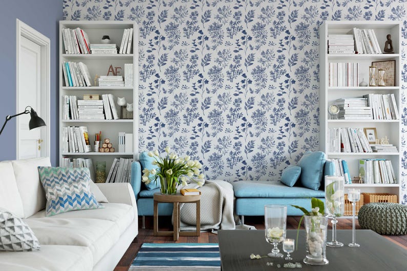 K&ouml;nnte beinhalten: Ein Wohnzimmer mit einem wei&szlig;en Sofa, blauen und wei&szlig;en Kissen mit Mustern, einem blauen Sofa und einem wei&szlig;en B&uuml;cherregal voller B&uuml;cher. Die Wand ist mit einer blauen und wei&szlig;en Blumentapete bedeckt.