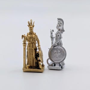 May include: Two small statues of a female figure, one in gold and one in silver. Both figures are wearing a crown and holding a staff and a shield. The shield has a design of a bird on it.