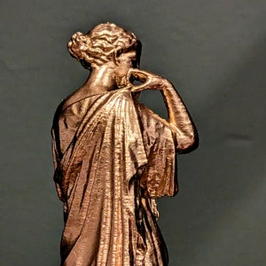 May include: A copper-colored statue of a woman wearing a flowing robe. The statue is posed with her head turned to the side and her hand resting on her chin.