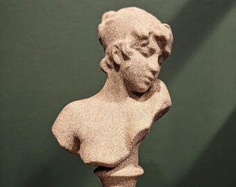 May include: A detailed bust sculpture of a person with curly hair, in a light beige color. The sculpture is on a pedestal base and is set against a dark green background. The bust is a classical art piece.