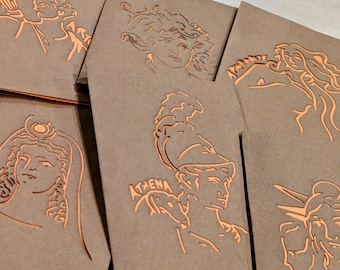 May include: A collection of kraft paper greeting cards with orange foil line art illustrations of Greek mythological figures. One card features the word "ATHENA". The cards are stacked and slightly fanned out.