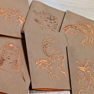May include: A collection of kraft paper greeting cards with orange foil line art illustrations of Greek mythological figures. One card features the word "ATHENA". The cards are stacked and slightly fanned out.