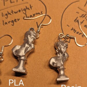This photo shows a closeup of a plastic PLA Sappho Bust earring next to a resin Sappho Bust earring.