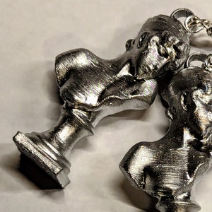 This photo shows a closeup of two silver-toned earrings featuring painted PLA charms of a Sappho bust and silver-plated stainless steel hooks. The earrings lie on a white background.