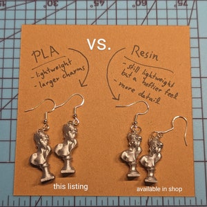 This photo shows a pair of plastic PLA Sappho Bust earrings next to a pair of resin Sappho Bust earrings. Both earrings lie on a brown kraft paper background. Text states that the resin earrings are heftier and more detailed.