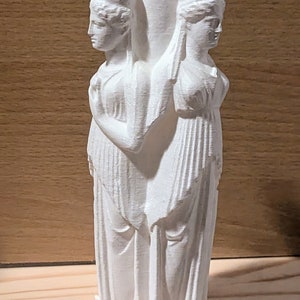 May include: A white, three-faced vase featuring classical female figures. The vase has a central cylindrical neck and two smaller openings, each adorned with a sculpted face. The figures are draped in flowing garments, creating a classical aesthetic. The vase is set against a wooden background.