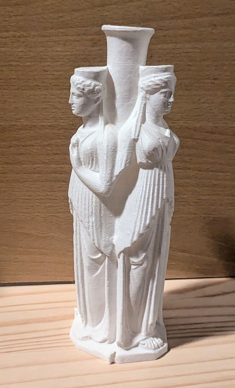 May include: A white, three-faced vase featuring classical female figures. The vase has a central cylindrical neck and two smaller openings, each adorned with a sculpted face. The figures are draped in flowing garments, creating a classical aesthetic. The vase is set against a wooden background.
