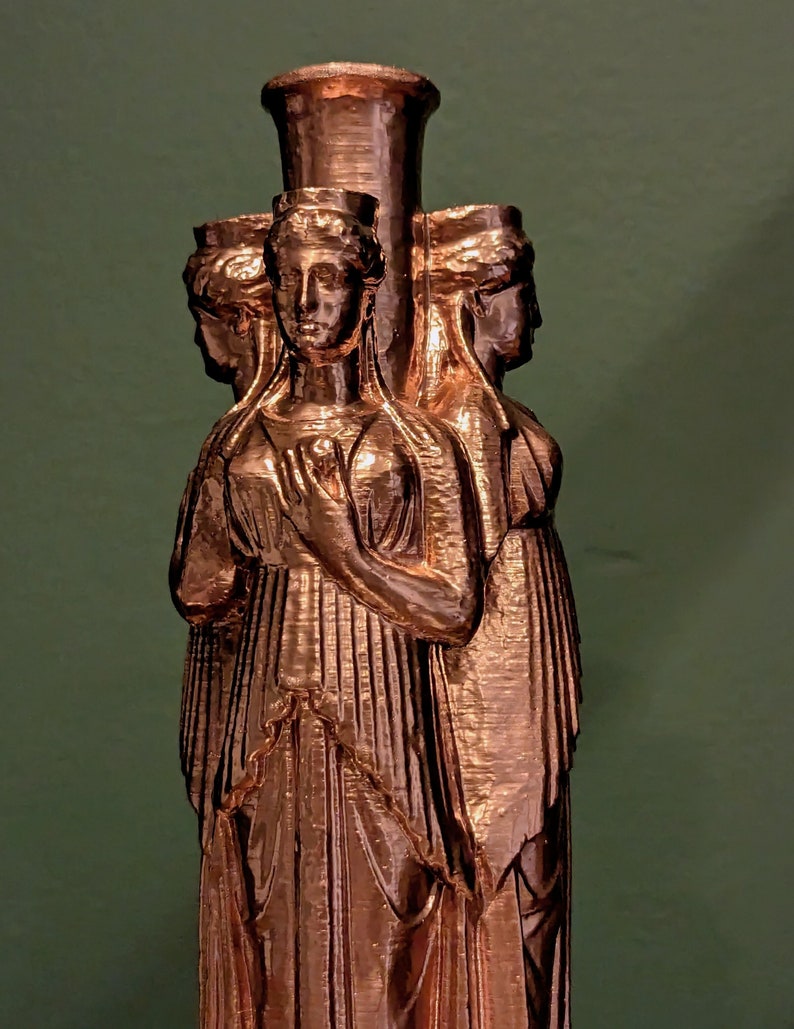 May include: A bronze-colored sculpture featuring three female figures with classical attire and hairstyles. The figures are arranged around a central column, with their faces visible. The sculpture has a textured surface and is set against a green background.