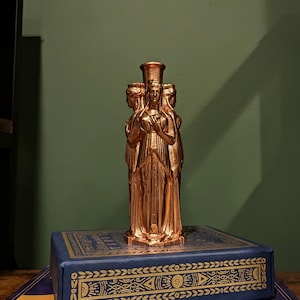This photo shows a bronze triple goddess Hecate statue. It sits on top of a blue and gold tarot deck box in front of a dark green background.