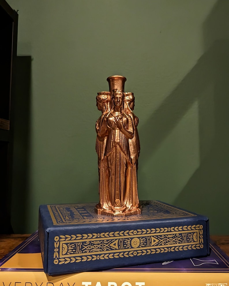 This photo shows a bronze triple goddess Hecate statue. It sits on top of a blue and gold tarot deck box in front of a dark green background.