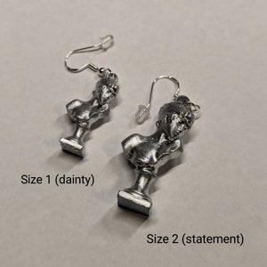 This photo shows a closeup of two sizes of silver-toned earrings featuring painted PLA charms of a Sappho bust and silver-plated stainless steel hooks 0 size one (dainty) and size 2 (statement). The earrings lie on a white background.