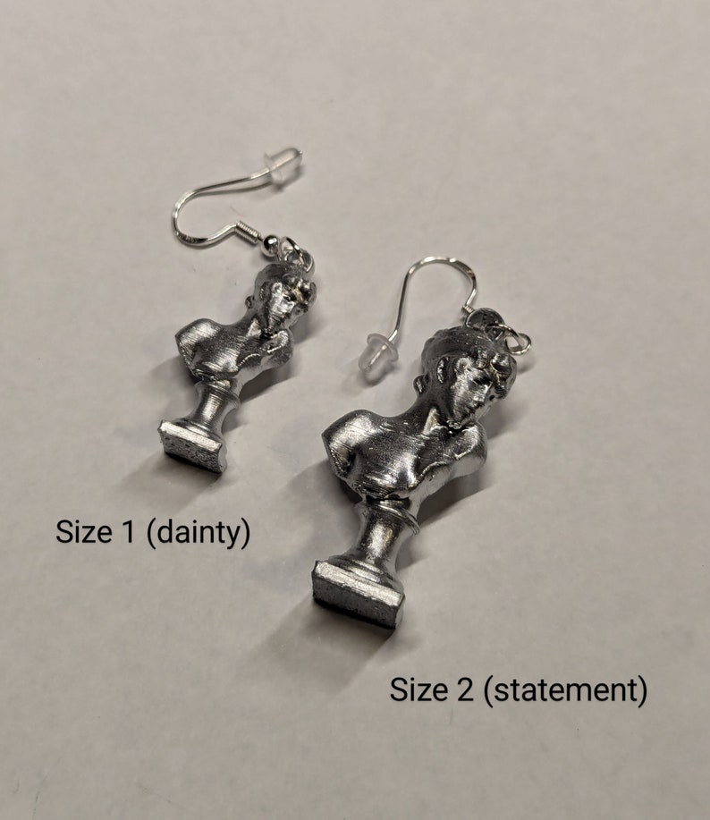 This photo shows a closeup of two sizes of silver-toned earrings featuring painted PLA charms of a Sappho bust and silver-plated stainless steel hooks 0 size one (dainty) and size 2 (statement). The earrings lie on a white background.