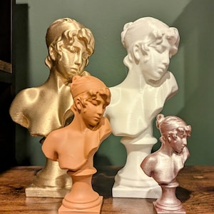 May include: Four bust sculptures of people with different hairstyles. The sculptures are made of a white, gold, and orange material. The sculptures are on a wooden surface.