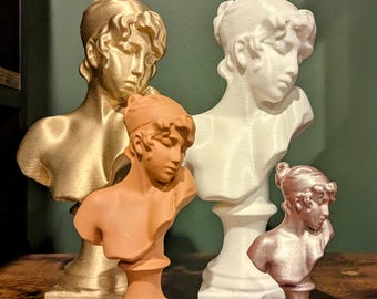 May include: Four bust sculptures of people with different hairstyles. The sculptures are made of a white, gold, and orange material. The sculptures are on a wooden surface.
