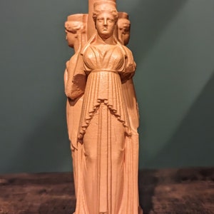 This photo shows a terracotta triple goddess Hecate statue. It sits on top of a dark wooden surface in front of a dark green background.
