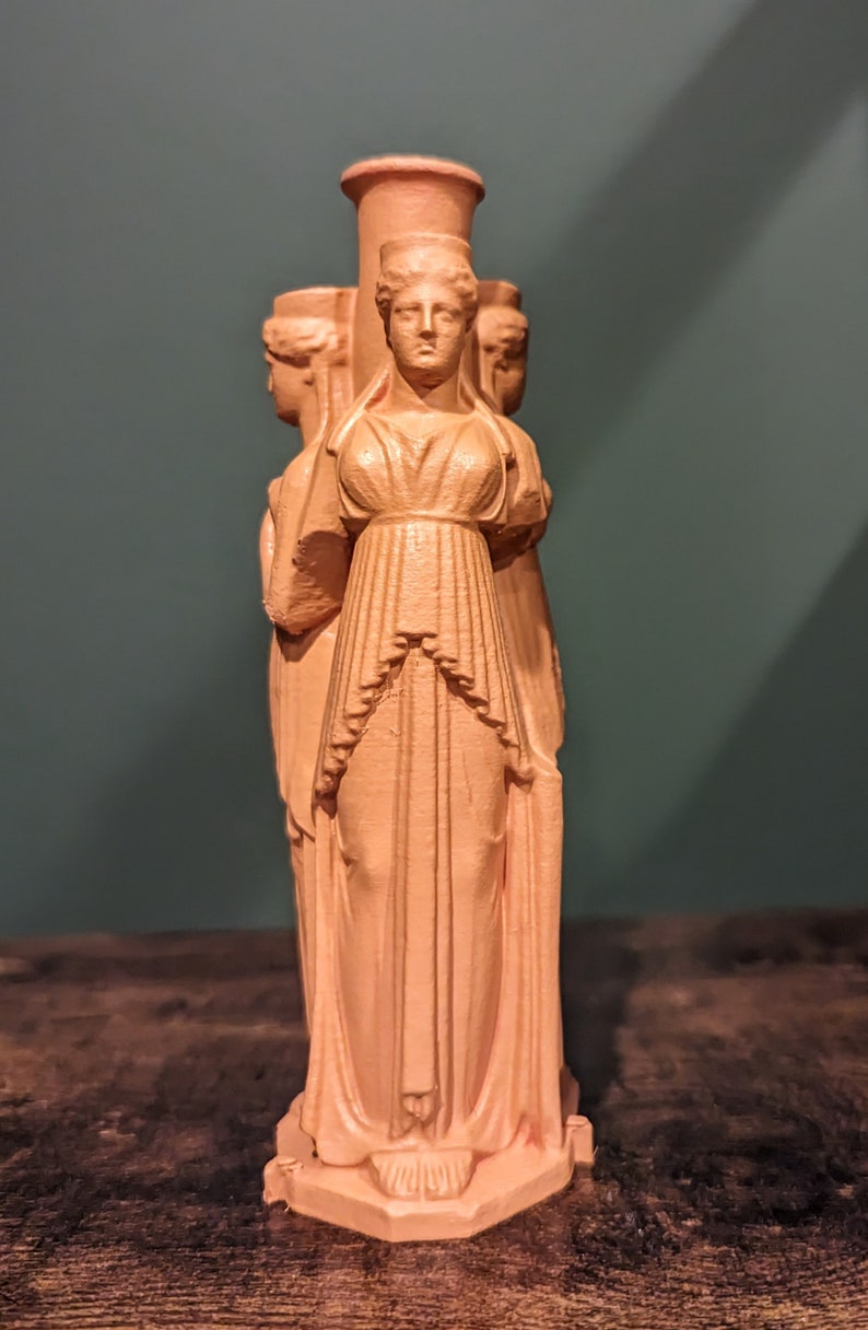 This photo shows a terracotta triple goddess Hecate statue. It sits on top of a dark wooden surface in front of a dark green background.