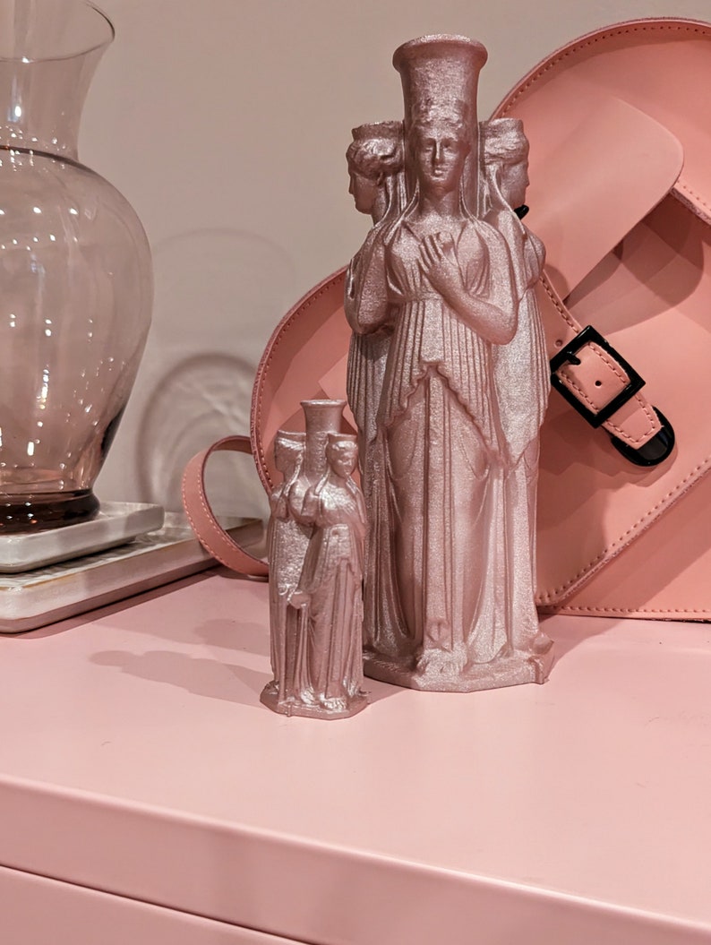 This photo shows two Hecate statues -one 9 inches tall and one 4 inches tall. Both are painted rose gold and sit on a pink cabinet in front of a pink, heart-shaped purse and a pink vase.