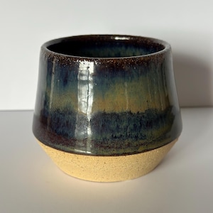May include: A ceramic cup with a wide, tapered body featuring a smooth, glossy glaze in shades of deep navy blue, forest green, and earthy brown. The lower portion of the cup remains unglazed, revealing a textured, light beige speckled clay base.