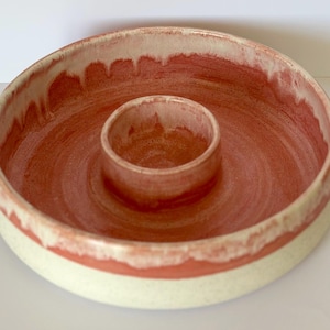 May include: A round ceramic serving dish with a central dipping bowl. The interior features a terracotta-coloured glaze with a drip pattern, while the exterior base is a speckled off-white. The rim shows a transition between the two colours.