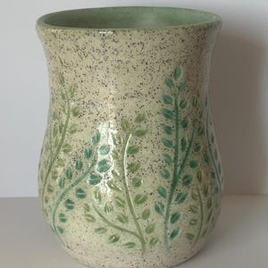 May include: A ceramic vase with a speckled cream-coloured glaze featuring embossed green leaf patterns. The vessel has a slightly flared rim and a rounded body, with the leaf designs wrapping around the exterior in shades of light and dark green.