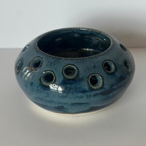 May include: A round, low-profile ceramic flower frog or vase with a glossy, deep blue glaze. The vessel features a wide, open centre and a series of evenly spaced, circular holes around the top shoulder, designed for arranging individual stems.