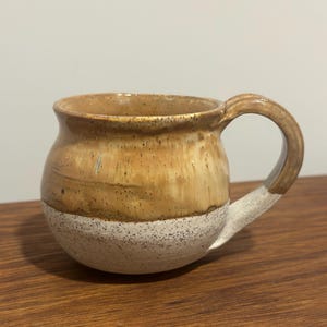 May include: A handmade ceramic mug with a rounded body and a large handle. The top half is a warm brown colour with a speckled texture, while the bottom half is a speckled white. The mug is designed for hot beverages.