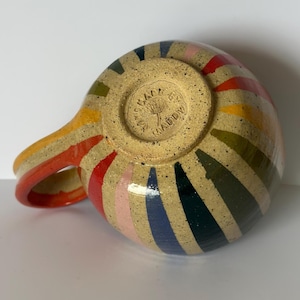 Pode incluir: Uma caneca de cerâmica salpicada com um padrão radial de listras multicoloridas em vermelho, amarelo, verde, azul e rosa. A base da caneca é estampada com o texto MADE BY MADDY e uma ilustração de dente-de-leão. A alça é curva e laranja.